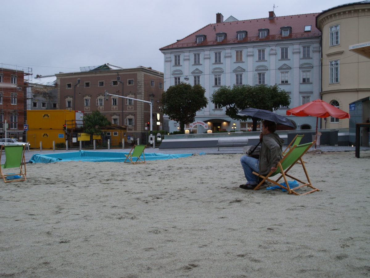 Dani a la platja artificial que van fer a Linz en una plaça amb el paraigua obert, plovent i tot desert