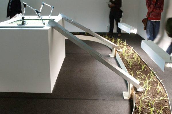 Instal·lació a una sala del museu, amb un carril pel de terra i plantes, uns robots i unes rampes